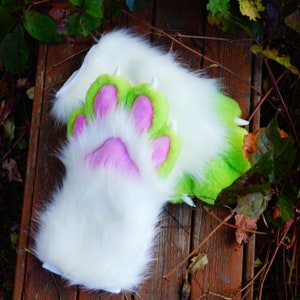 May include: A pair of white furry paws with pink and green paw pads and claws. The paws are lying on a wooden surface.