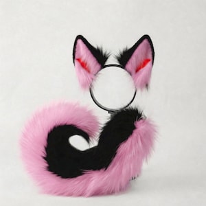 May include: A pink and black faux fur tail and matching cat ear headband set. The ears are black with pink and red accents. The tail is pink with a black stripe. The set is designed for cosplay or costume use.