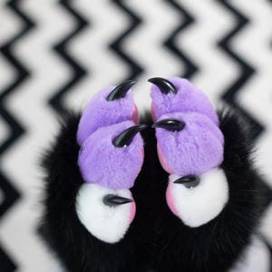 May include: Close-up of plush monster paws. The paws are lavender with black claws and pink accents. The fur around the paws is black and white. The background is a black and white zig-zag pattern.