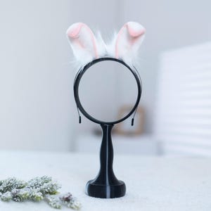 May include: A white bunny ear headband with pink tips on a black stand. The headband is made of soft, fluffy material and is perfect for adding a touch of whimsy to any outfit.
