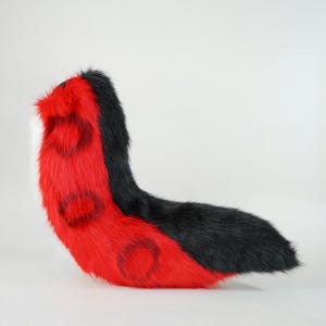 May include: A long, curved faux fur tail featuring a vibrant red base color with distinct black circular markings along the side. The top section of the tail is solid black, while the very tip of the tail displays a small patch of white fur.