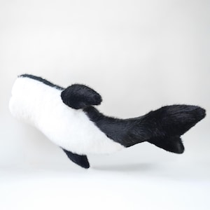 May include: A plush toy whale in black and white. The toy has a white body with black accents on the fins, tail, and top of the head. The whale is lying on its side against a white background.