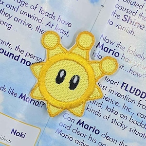 May include: Embroidered yellow sun patch with a smiling face, featuring a circular design with radiating points. The patch is placed on a colorful page with text and illustrations, likely from a video game guide.