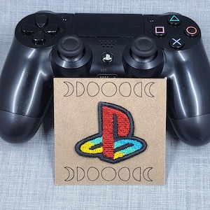 May include: A brown card with a white PlayStation logo embroidered on it. The card is sitting on top of a black PlayStation controller.