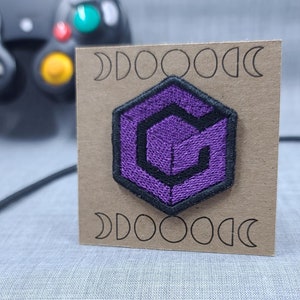May include: Purple and black embroidered patch of the GameCube logo on a brown card with the text "DOOOOOC" on the top and bottom.