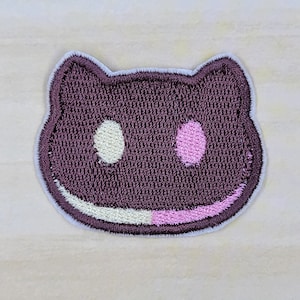 May include: A purple embroidered patch shaped like a cat face with white and pink details. The cat has a white eye on the left and a pink eye on the right. The mouth is a white and pink smile.