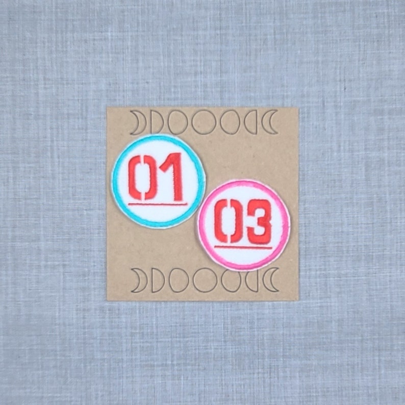 Puede incluir: Dos parches bordados con fondo blanco y n&uacute;meros rojos. El primer parche es azul y dice "01" y el segundo parche es rosa y dice "03".