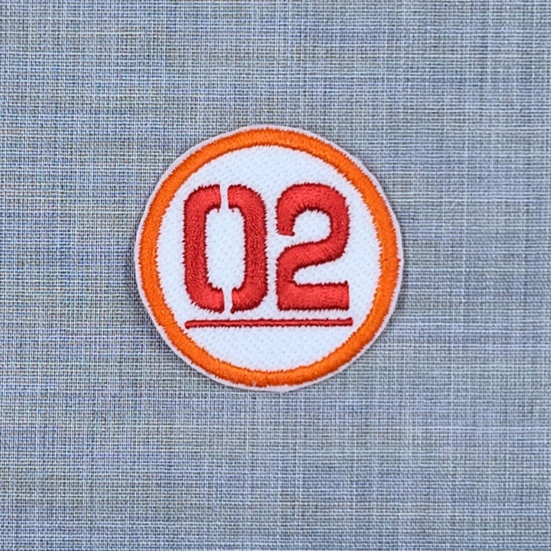 Puede incluir: Un c&iacute;rculo blanco con borde naranja y el n&uacute;mero "02" bordado en rojo.