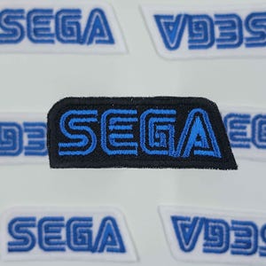 May include: A black and blue embroidered patch with the word "SEGA" in a stylized font.