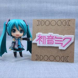 May include: A white patch with pink embroidered text that reads "初音ミク" on a brown background with a white moon design. A turquoise-haired anime figurine is standing in front of the patch.