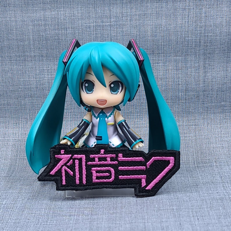 May include: A turquoise blue anime character with long hair and a pink and black patch that says "初音ミク" in Japanese.