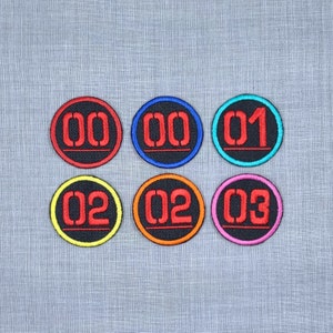 May include: Six embroidered iron-on patches with black backgrounds and red numbers. The numbers are 00, 01, 02, and 03. The patches have different colored borders: red, blue, turquoise, yellow, orange, and pink.