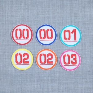 May include: Six round embroidered iron-on patches with white backgrounds and red numbers. The numbers are 00, 01, 02, and 03. The patches have different colored borders: red, blue, turquoise, yellow, orange, and pink.