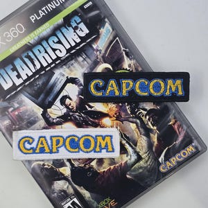 May include: Two embroidered patches with the word "CAPCOM" in yellow letters with a blue outline. The patches are on a black and white background. The background is a video game case for the Xbox 360 game "Dead Rising".