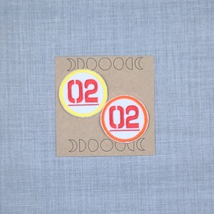 Puede incluir: Dos parches bordados para planchar con el n&uacute;mero "02" en rojo y blanco. El parche de la izquierda tiene un c&iacute;rculo exterior amarillo y el parche de la derecha tiene un c&iacute;rculo exterior naranja.