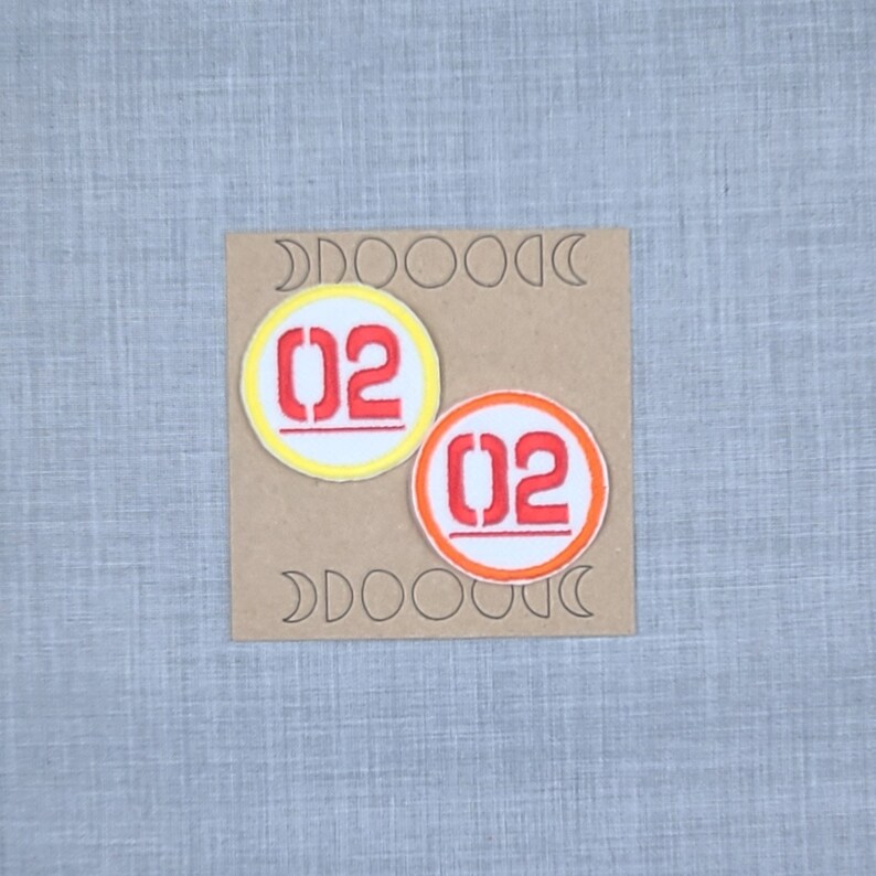 Puede incluir: Dos parches bordados para planchar con el n&uacute;mero "02" en rojo y blanco. El parche de la izquierda tiene un c&iacute;rculo exterior amarillo y el parche de la derecha tiene un c&iacute;rculo exterior naranja.