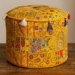 Op de afbeelding: Een ronde, gele poef met ingewikkeld patchwork en geborduurde bloemmotieven. De poef heeft een cilindrische vorm met decoratieve kwastjes in verschillende kleuren. Het ontwerp bevat een mix van patronen en texturen, wat een bohemien esthetiek creëert.