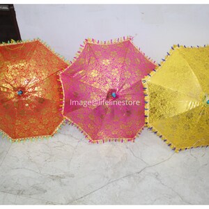 Puede incluir: Tres sombrillas decorativas con tela dorada, rosa y amarilla. Las sombrillas tienen un borde con flecos y están cerradas.