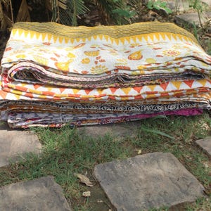 May include: A stack of colourful kantha quilts, folded and arranged on a stone path. The quilts are made of cotton fabric and feature a variety of patterns and colours, including yellow, orange, red, pink, and blue.