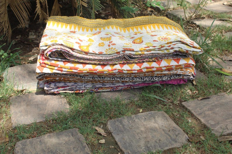 May include: A stack of colourful kantha quilts, folded and arranged on a stone path. The quilts are made of cotton fabric and feature a variety of patterns and colours, including yellow, orange, red, pink, and blue.
