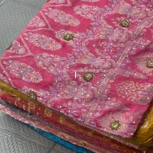 Pode incluir: Uma pilha de peças de tecido coloridas, apresentando um tecido rosa vibrante com bordados florais intrincados e enfeites de lantejoulas. Outros tecidos na pilha incluem tons de amarelo, vermelho e azul. Os têxteis parecem estar dobrados e prontos para uso.