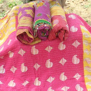 May include: A pink and yellow kantha quilt with a paisley pattern. The quilt is folded and rolled up, with the ends of the fabric visible.