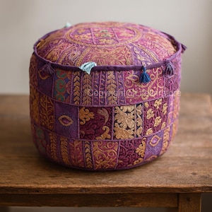 Op de afbeelding: Een ronde, paarse poef met ingewikkelde gouden borduursels en patchwork-ontwerp. De poef heeft een gewatteerde boven- en zijkanten, met kleine blauwe kwastjes. Het ontwerp bevat bloemen- en geometrische patronen, wat een bohemien esthetiek creëert.