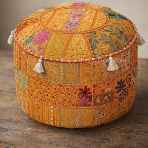 Puede incluir: Puf redondo amarillo con diseño patchwork. El puf presenta varios patrones florales y geométricos en tonos naranja, rosa y verde. Borlas blancas adornan los lados. El puf está colocado sobre una superficie de madera.
