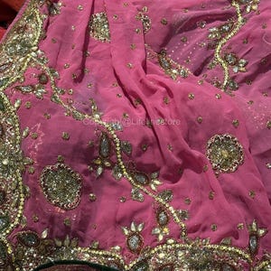 May include: A pink textile with elaborate gold and silver detailing. The fabric is embellished with detailed embroidery, sequins, and beadwork, creating a luxurious and ornate appearance. The design features floral and geometric patterns.