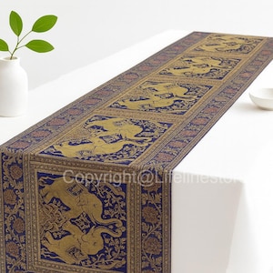 May include: A rectangular table runner with a navy blue border and gold elephant design. The runner is placed on a white tablecloth. A small white vase with a green plant and a white bowl are also on the table.