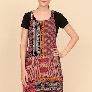 Puede incluir: Un delantal patchwork con una variedad de patrones y colores, incluyendo rojo, negro y dorado. El delantal tiene una parte superior de babero y un bolsillo grande. Se lleva sobre una camiseta negra.