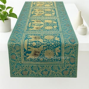 Puede incluir: Camino de mesa turquesa y dorado con un estampado de elefantes y flores. El camino de mesa rectangular presenta un diseño repetido de elefantes y flores doradas sobre un fondo turquesa. El camino de mesa es de tela texturizada.
