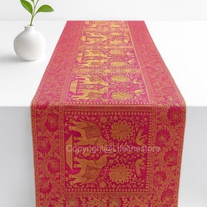 Peut inclure: Chemin de table rose vif avec un motif doré d'éléphants et de fleurs. Le chemin de table rectangulaire est présenté sur une surface blanche, mettant en valeur le motif complexe. Un vase blanc avec une plante verte est en arrière-plan.