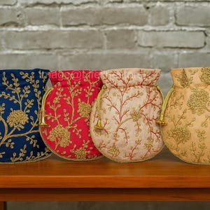 Könnte beinhalten: Vier bestickte Kordelzugbeutel in Marineblau, Rot, Creme und Gold. Jeder Beutel hat ein Blumendesign mit Goldfaden und einen passenden Kordelzugverschluss. Die Beutel werden auf einem Holztisch ausgestellt.