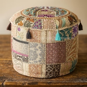 May include: A round, patchwork pouf with a flat top and cylindrical shape. The pouf features a variety of patterned fabric squares in shades of beige, brown, purple, and teal. Small tassels in various colors adorn the sides, adding a decorative touch.