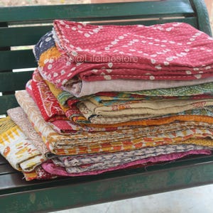 May include: A stack of colourful kantha quilts, made from recycled saris, are arranged on a green wooden bench. The quilts are folded and layered, showcasing a variety of patterns and textures.