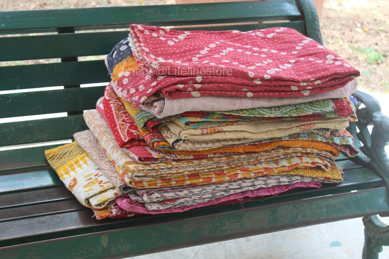 May include: A stack of colourful kantha quilts, made from recycled saris, are arranged on a green wooden bench. The quilts are folded and layered, showcasing a variety of patterns and textures.