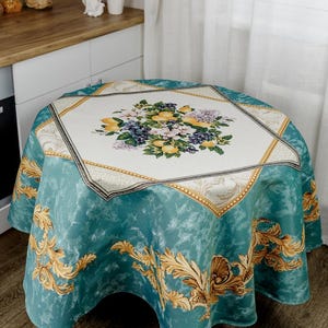 以下が含まれることがあります： 金色と白のフローラルデザインが施された丸いターコイズブルーのテーブルクロス。テーブルクロスの中央には、レモン、ブドウ、花をモチーフにしたデザインが施されています。テーブルクロスの縁は、金色と白のパターンで飾られています。