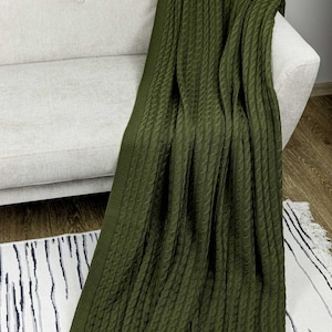 Könnte beinhalten: Eine olivgrüne Zopfstrickdecke, die über dem Arm eines hellgrauen Sofas drapiert ist. Die Decke hat ein strukturiertes Muster und ist aus einem weichen Material gefertigt. Die Decke liegt auf einem schwarz-weiß gestreiften Teppich.