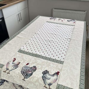 Könnte beinhalten: Eine beige Tischdecke mit einem sich wiederholenden Blumenmuster und einem zentralen Tischläufer. Die Tischdecke hat einen Rand mit einem grün-weißen Design. Die Ecken und der Läufer sind mit Bildern von Hühnern verziert.