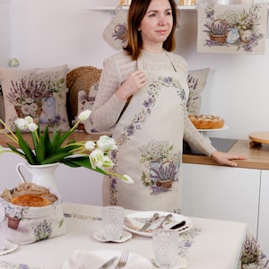 May include: A woman wearing a beige apron with a lavender floral design. The apron, tablecloth, and other kitchen textiles feature a matching lavender and floral theme. A white pitcher of flowers and a loaf of bread are on the table.