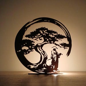 May include: A black laser-cut silhouette of a bonsai tree and a figure of a person in traditional attire, set within a circular frame. The artwork is placed on a wooden surface, illuminated by a soft light.