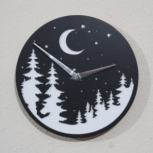 May include: A black and white clock with a moon and stars design. The clock face features a silhouette of a forest with trees and a crescent moon. The clock hands are silver.