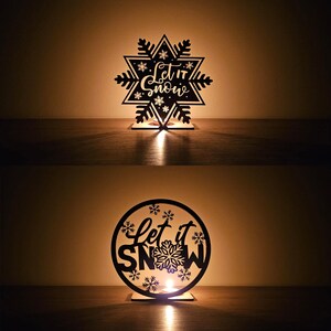 以下が含まれることがあります： 「Let it Snow」の文字が切り抜かれた、黒い木製のキャンドルホルダーが2つ。1つは雪の結晶の形、もう1つは丸型です。どちらも中に小さなキャンドルが灯されており、温かい光を放っています。