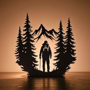 Può includere: Una silhouette nera di un escursionista con zaino e bastoncino, incorniciata da pini e montagne. Il design è tagliato da un unico pezzo di materiale, creando un effetto a strati. Lo sfondo è di colore ambra caldo.