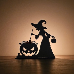 May include: A black silhouette of a witch stirring a cauldron with a jack-o'-lantern face. The witch is wearing a pointed hat and holding a small bag. The Halloween-themed decoration is set on a wooden surface.