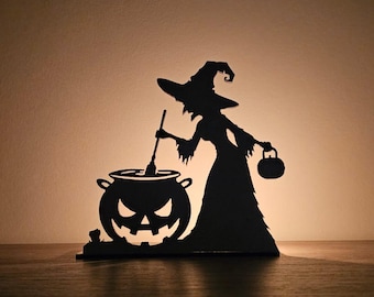 Puede incluir: Silueta negra de una bruja removiendo un caldero con una cara de calabaza. La bruja lleva un sombrero puntiagudo y sostiene una pequeña bolsa. Decoración temática de Halloween, colocada sobre una superficie de madera.