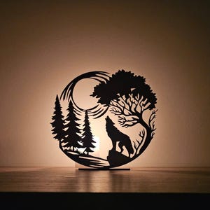 May include: Black silhouette of a howling wolf, trees, and moon in a circular design. The artwork is cut from a single piece of material and is placed on a wooden surface. The background is a warm, neutral color.