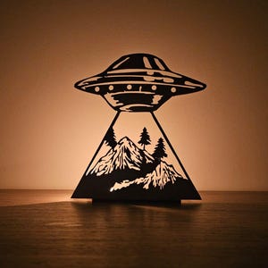 Può includere: Lampada a forma di UFO tagliata al laser, nera, con una scena di montagna e alberi. La lampada presenta un design a forma di disco volante sopra una base triangolare con dettagliate silhouette di montagne e alberi. La lampada è illuminata, proiettando un bagliore caldo.