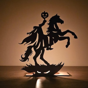Puede incluir: Silueta negra del Jinete sin Cabeza montando un caballo encabritado, sosteniendo una linterna de calabaza. El caballo y el jinete están sobre una base de hierba con una cara de linterna de calabaza tallada. Decoración de Halloween de madera.